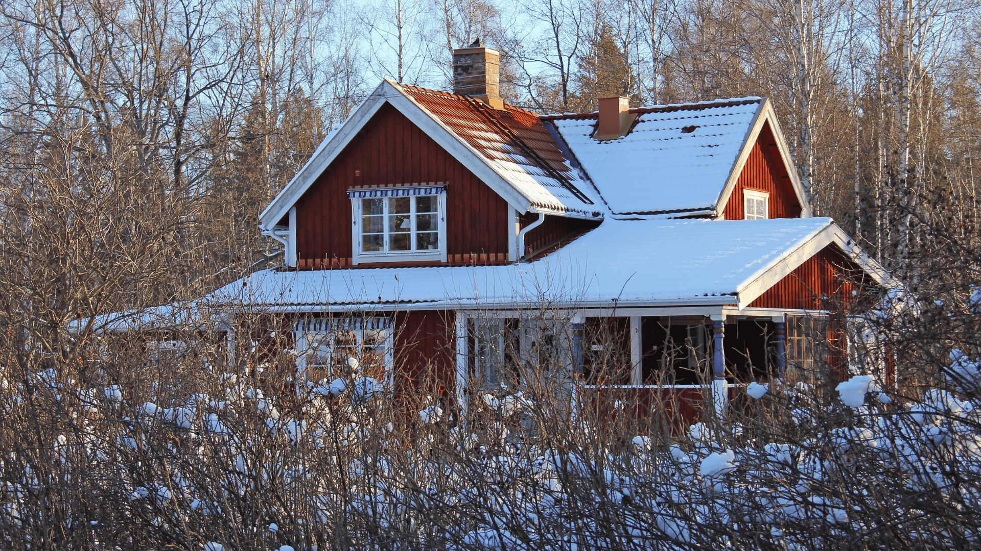 Ett rött trähus med snö på taket, omgivet av träd och buskar, som symboliserar hur man kan vinterförbereda huset inför kylan.
